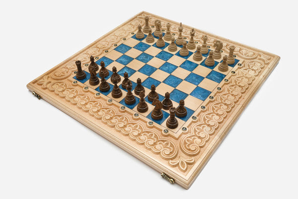Large folding chess board in pale wood with blue epoxy squares, a carved fleur-de-lis border and wooden pieces set up for play