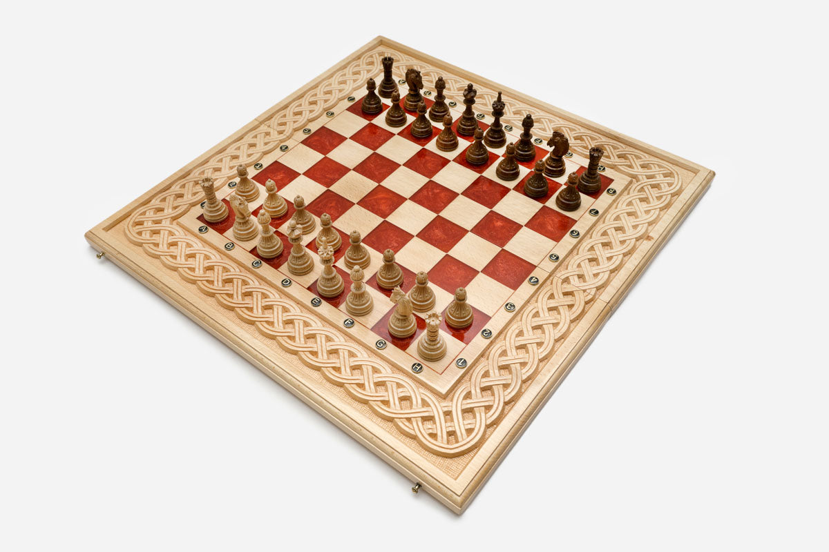 Large folding chess board in pale wood with red epoxy squares, a carved Celtic-knot border and wooden pieces set up for play