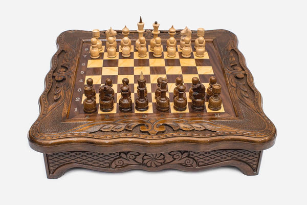 Compact carved wooden chess set with scalloped frame and wooden pieces set up on the honey-walnut board