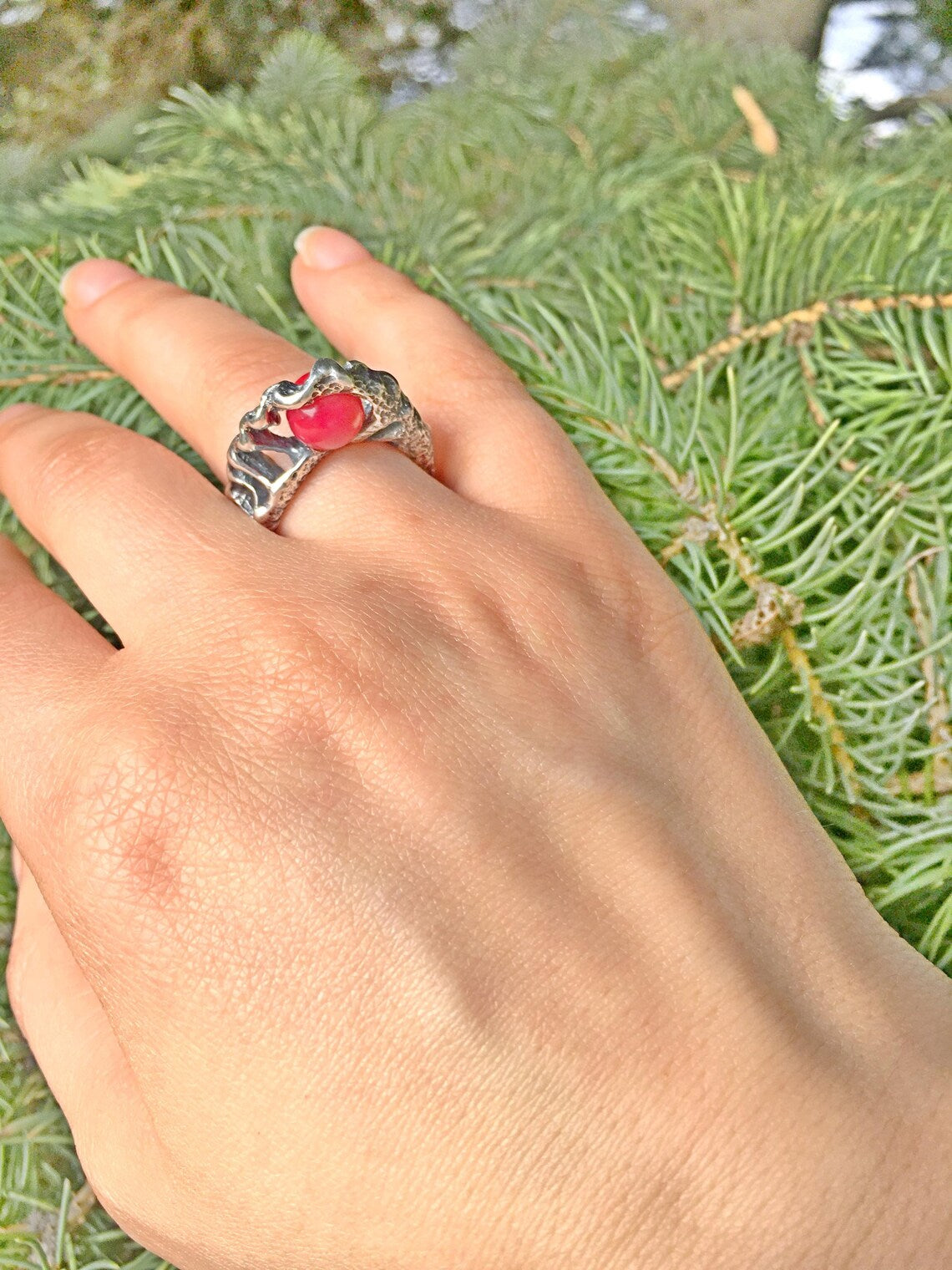 Hand wearing a silver ring with a red stone against a green foliage background