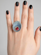 Silver ring with a red gemstone on a hand with black nail polish against a gray background