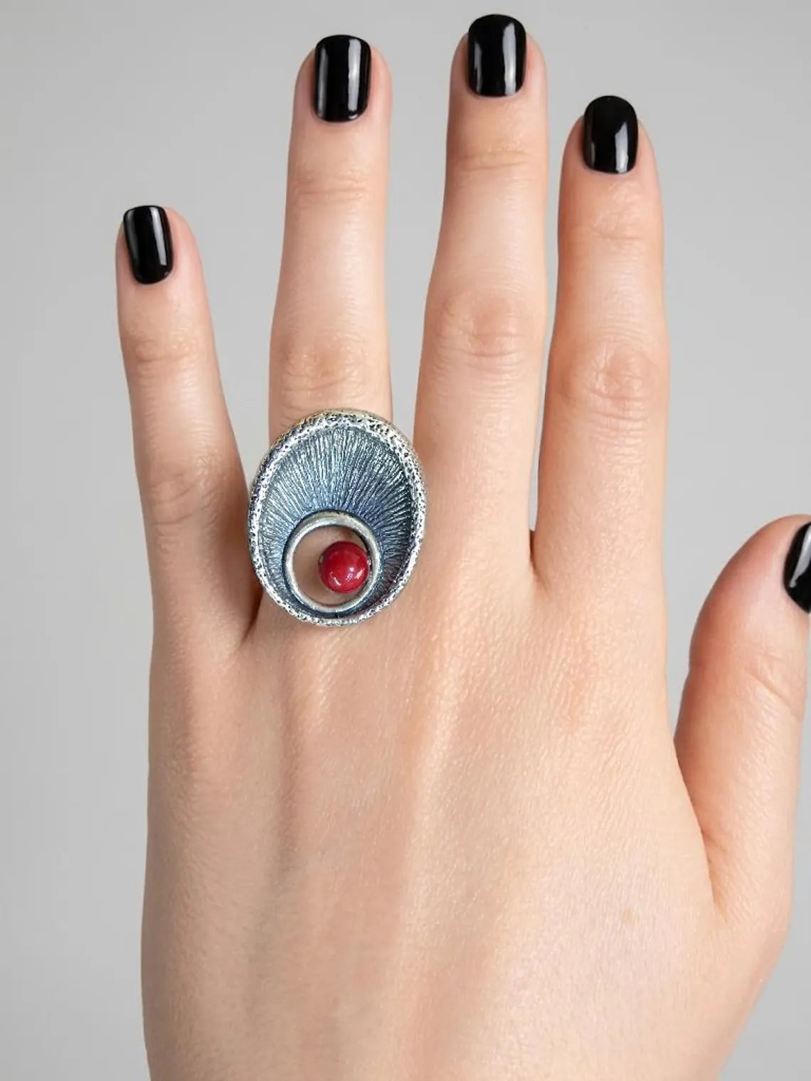 Silver ring with a red gemstone on a hand with black nail polish against a gray background